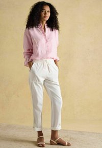 Cămașă cu dungi roz și albe, cu nasturi și manșete cu volane, combinată cu pantaloni albi cu talie elastică și sandale joase de culoare deschisă. Design minimalist.