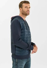 Chaqueta acolchada navy con cremallera y mangas de una tela navy contrastante, puños elásticos y un forro de capucha rojo y blanco. Cuenta con bolsillos laterales.