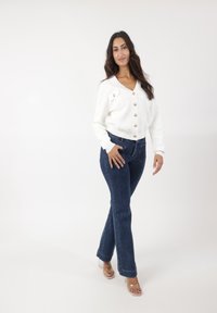 Witte geweven cardigan met gouden knopen, gecombineerd met donkerblauwe flared jeans. Model draagt doorzichtige haksandalen en staat tegen een effen achtergrond.