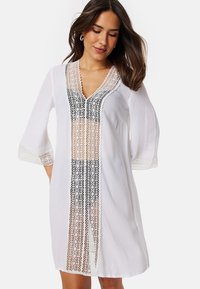 Copricostume bianco con maniche a tre quarti, caratterizzato da un design in pizzo lungo la parte anteriore e intricati dettagli in pizzo sulle maniche. Tessuto testurizzato.