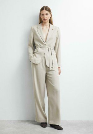 Femme aux longs cheveux portant un blazer beige ceinturé et un pantalon à jambes larges, debout contre un mur blanc les mains dans les poches.