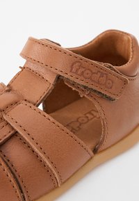 Smeđi kožni sandal s zatvorenim prstima, višestrukim trakama i čičak zatvaranjem. Ima šivane ukrase i svijetlosmeđi potplat.