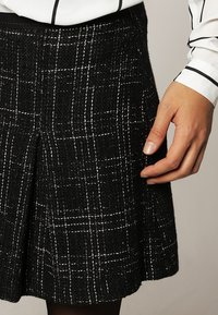 Gonna a scacchi nera realizzata in materiale testurizzato, con linee bianche, pieghe e una vita aderente. Una mano è posizionata sulla gonna.