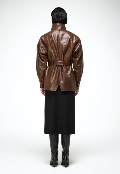 Personne portant une veste en cuir marron ceinturée, une jupe noire mi-longue et des bottes à talons noires, vue de dos sur un fond blanc.