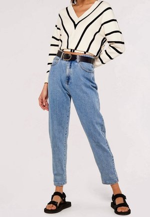 Femme portant un pull en V rayé noir et blanc, un jean bleu taille haute avec une ceinture noire, et des sandales noires à bouts ouverts.