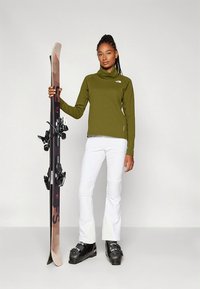 Nő olívazöld felsőben, fehér sí nadrágban, fekete síbakancsban, egy pár függőlegesen tartott síléccel egy egyszínű fehér háttér el�őtt.