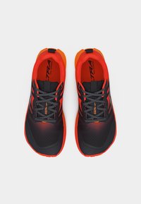 Ζευγάρι μαύρα και πορτοκαλί παπούτσια τρεξίματος σε μονοπάτι με κορδόνια με σχέδιο, ορατά από πάνω σε λευκό φόντο.