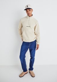 Sudadera de forro polar beige con media cremallera, que cuenta con un bolsillo frontal y un estampado de "Originals Studio". Combinada con unos vaqueros azules y zapatillas beige.