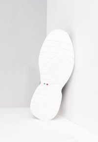 Semelle de sneaker blanche avec motif de grip texturé, présentant un accent vert, blanc et rouge près du talon, placée sur un fond clair.