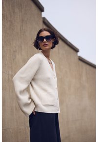 Cardigan a coste color crema con scollo a V, chiusura a bottoni e tasche laterali. Abbinato a pantaloni neri, indossato con occhiali da sole neri oversize.