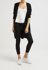 Cardigan lungo nero sopra una canotta bianca con bordo a pompon, abbinato a jeans skinny neri e sneakers bianche. Design semplice e stile aderente.