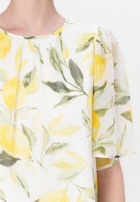 Blomsterbluse i lett stoff, med gule sitroner og grønne blader på en hvit bakgrunn. Myke, gjennomsiktige ermer med rund hals.