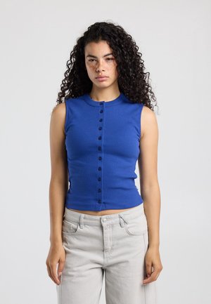 Vrouw met krullend haar en sproeten die een mouwloos blauw topje met knopen en lichtgrijze broek draagt, staand tegen een effen witte achtergrond.