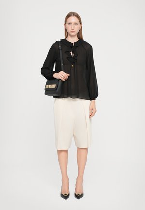 Black sheer blouse with ruffle accents, cream tailored shorts, and black pointed-toe heels. Model holds a black bag with gold lettering.