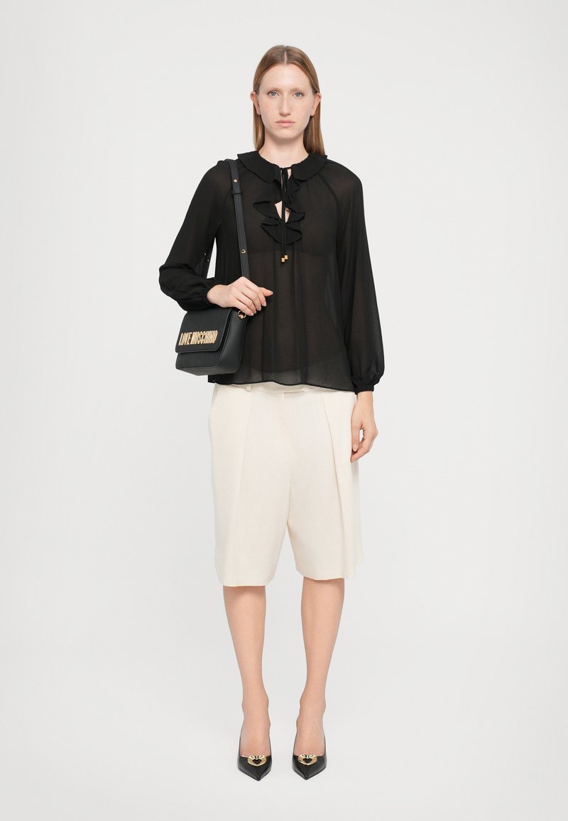 Bluza neagră din material transparent cu accente volan, pantaloni scurți crem, adaptați, și pantofi cu vârf ascuțit negri. Modelul ține o geantă neagră cu litere aurii.