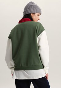 Zelená mikina s bílými rukávy, žebrovanými manžetami a červeným límcem. Má volný střih a pod ní je vrstvená košile.