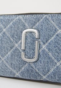 Sinine trapetsiline denimkangas valgete õmblustega ja hõbedase üksteisega seotud "J" logo detailiga, nähtav ülemine tõmblukk.