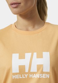 Camiseta de algodón naranja con escote redondo, que presenta un gran logo blanco "HH" y "HELLY HANSEN" impreso debajo en letras negritas.