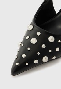 Chaussure en cuir noir à bout pointu, ornée de clous métalliques argentés de différentes tailles, avec un design élégant et ouvert.