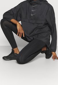 Zwart Nike windbreaker met een capuchon, voorzien van een subtiele glans en geborduurde logo's. Gecombineerd met zwarte leggings en getextureerde sportschoenen.