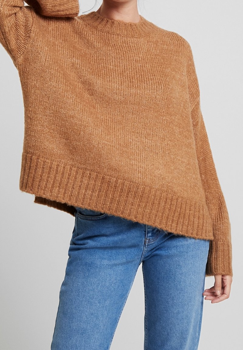 Persoon in een losse, geribde, camel-kleurige trui en blauwe jeans, die één arm omhoog houdt tegen een effen lichte achtergrond.