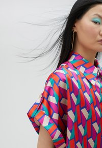 Colorful short-sleeve shirt made of satin fabric featuring a geometric pattern in pink, orange, white, and teal with button accents.