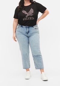 Černé tričko s nápisem "DREAMER" a designem orla v metalických akcentacích, spárované se světle modrými džíny s vysokým pasem a bílými teniskami.