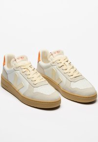 Zapatillas beige con suelas de goma, cuellos acolchados y cajas de dedos perforadas. Presentan acentos naranjas y un acabado texturizado en las superposiciones de ante.