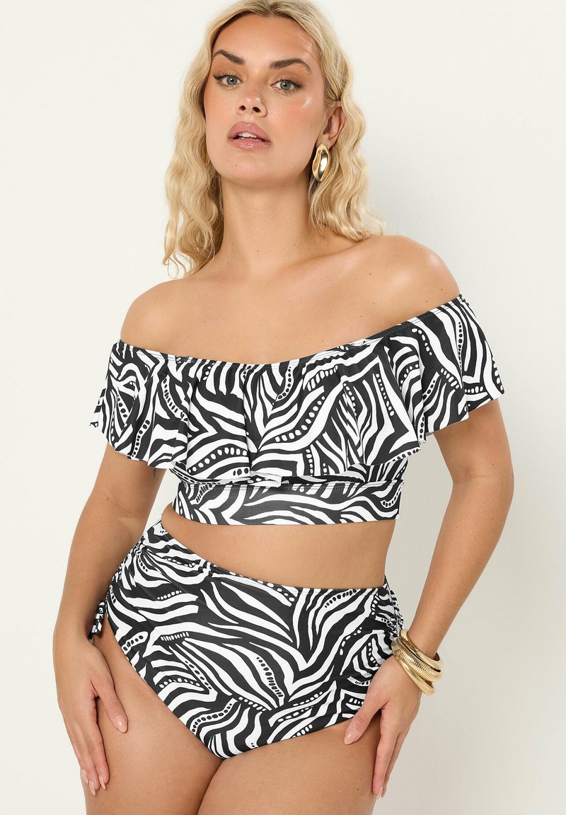 Vrouw die een zwart-witte zebra-patroon off-shoulder cropped top met ruches draagt en bijpassende hooggesneden bikinibroekjes, gouden oorringen en armbanden.
