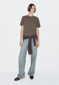 T-shirt in cotone marrone con maniche corte, abbinato a jeans di denim larghi e blu chiaro, e a un maglione scuro in maglia legato in vita.