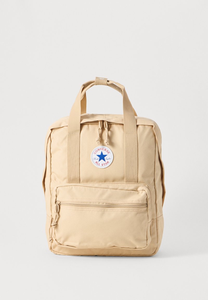 Beige tyg ryggsäck med rektangulär form, dubbla handtag upptill, ficka med dragkedja fram och en rund Converse All Star-logotyp.