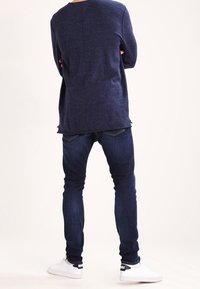 Person som står med armarna korsade, iförd en marinblå tröja, mörkblå jeans och vita Nike-sneakers, vänd bort från kameran mot en enfärgad bakgrund.