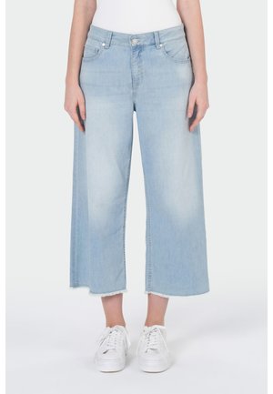 Personne portant un jean court larges jambes bleu clair avec ourlets bruts et baskets blanches, debout devant un fond uni.