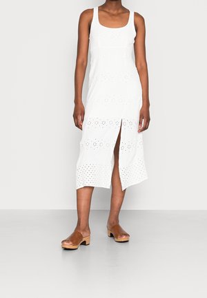 Femme portant une robe midi blanche sans manches avec un motif œillet et une fente à l'avant, associée à des sandales ouvertes marron à talons en bois.