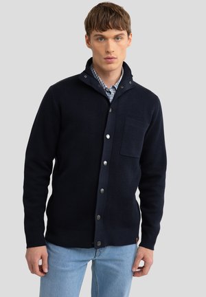 Marineblaue Strickjacke mit strukturiertem Finish, knöpfbarem Frontbereich und einer einzelnen Brusttasche. Sie verfügt über einen Kragen und silberfarbene Knöpfe.