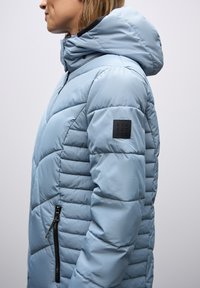 Giacca imbottita azzurro chiaro con cappuccio, dotata di design trapuntato, tasca laterale con zip e una patch nera con logo sulla manica.