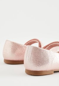 Paire de chaussures plates roses scintillantes et texturées avec semelles marron sur une surface blanche vue du côté du talon.