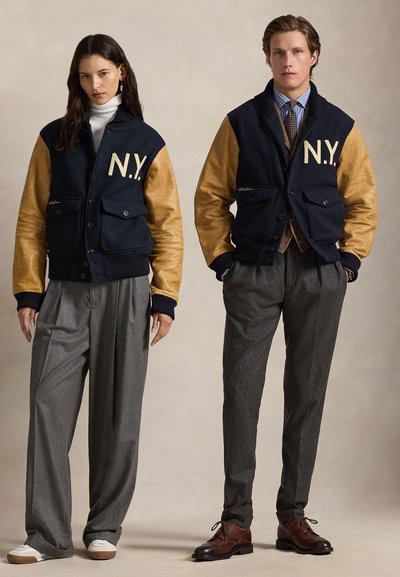Giubbotti varsity in misto lana blu navy con maniche in pelle beige, grande scritta "N.Y.", indossabili sopra un dolcevita bianco e un completo marrone, abbinati a pantaloni grigi.