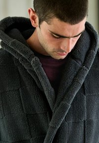 Mörkgrå, mjuk fleecebadrock med huva, som har en quiltad textur och krage. Bärs över en mörk skjorta, vilket visar på en avslappnad passform och mjukhet.