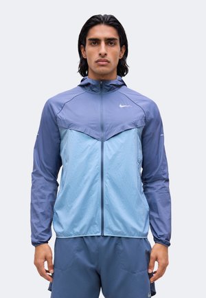 Homme aux cheveux noirs mi-longs portant une veste zippée Nike bicolore bleue et un short de sport assorti sur un fond uni.