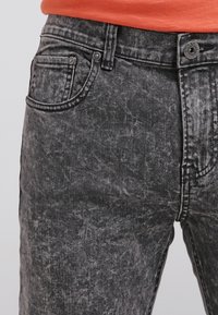 Černé džíny z acid-wash denimu s texturovaným povrchem, které mají tradiční kapsy a kovové zapínání na knoflík v pase.