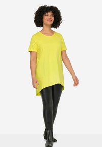 Tunic gialla a maniche corte con orlo curvo, abbinata a leggings neri e stivali alla caviglia con rifiniture a borchie. Tessuto morbido e leggero.