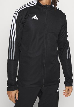 Mężczyzna ubrany w czarny dres Adidas z białymi paskami na rękawach oraz logo Adidas na piersi i spodniach, stojący na jednolitym tle.
