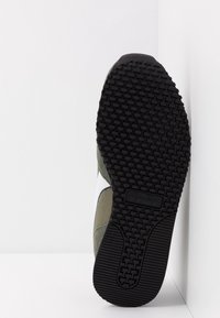 Zapatilla deportiva con suela de goma negra, parte superior de ante en verde y blanco, detalles con textura y diseño de bajo perfil.