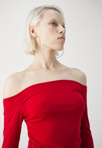 Top roșu, fără umeri, fabricat dintr-un material elastic, cu mâneci lungi și detalii pliate pe laterale.