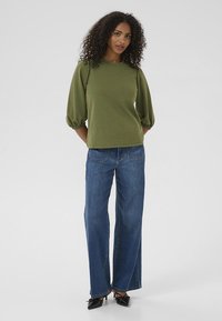 Olivengrøn tekstureret top med pufærmer, parret med brede blå jeans og sorte spidse hæle. Enkel og struktureret design.