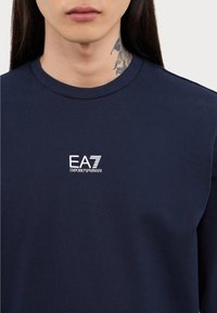 Tumši zila garām piedurknēm t-krekls, izgatavots no kokvilnas, ar baltu EA7 Emporio Armani logotipu uz krūtīm un arapa izgriezumu.