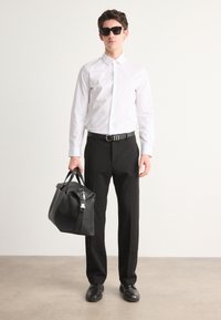 White collared shirt, black trousers, black belt, and black shoes. Carrying a textured black bag with a silver hardware accent. Black sunglasses.