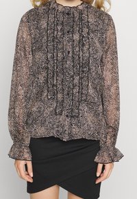 Blusa a maniche lunghe nera e beige a fantasia, con polsini arricciati e dettagli al collo raccolto, abbinata a una gonna avvolgente nera.