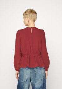 Haut peplum bordeaux avec de longues manches bouffantes, tissu texturé, taille froncée et un détail en forme de trou de serrure au dos. Associé à un jean bleu.
